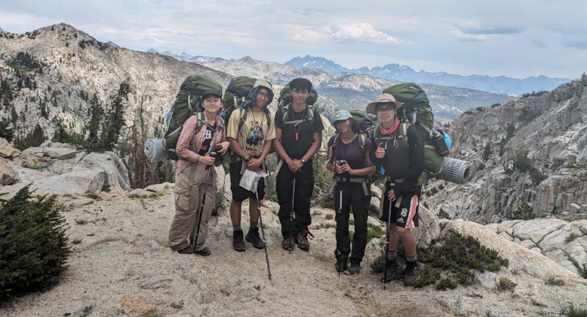 Five people wearing backpacks stand on a rocky overlook and smile for the photo. Behind them are the vast high sierra. 